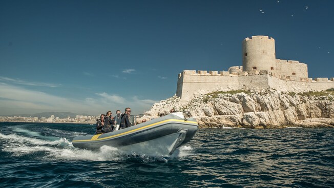 Guests wearing wetsuits traveling in a zodiac boat in front of a fort on a cliff