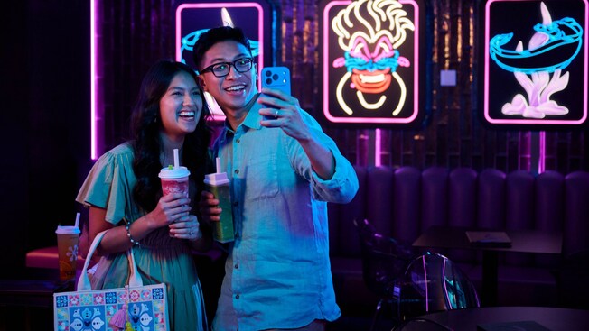 Two Guests enjoying beverages in a lounge surrounded by illuminated artwork at Bewitching Boba and Brews on the Disney Adventure cruise ship