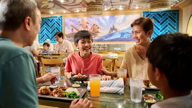 Uma família de quatro pessoas saboreando uma refeição no Gramma Tala's Kitchen, no navio de cruzeiro Disney Adventure Uma família de quatro pessoas saboreando uma refeição no Gramma Tala's Kitchen, no navio de cruzeiro Disney Adventure