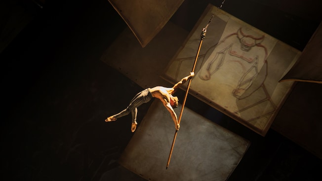 Acrobata aéreo atravessa o ar segurando um lápis perto de um rascunho do Chernabog