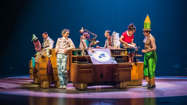 Um grupo de pessoas perto de pranchetas de desenho enquanto Julie encara Mister Pencil