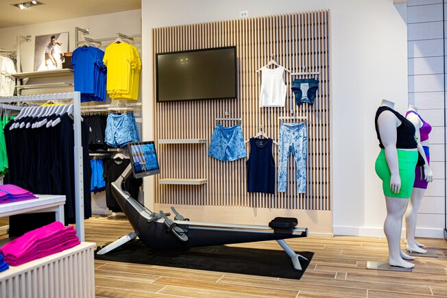 El interior de una tienda de Fabletics, con una máquina de remo, 2 maniquíes con ropa deportiva y estantes repletos de ropa deportiva