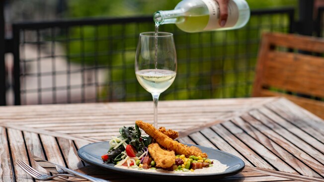 At an outdoor table, a hand pours wine into a glass, which is next to a tempting plate of catfish and succotash