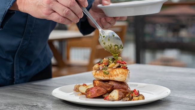 A man’s hand spoons salsa verde over a piece of Mahi, which rests on a bed of potatoes
