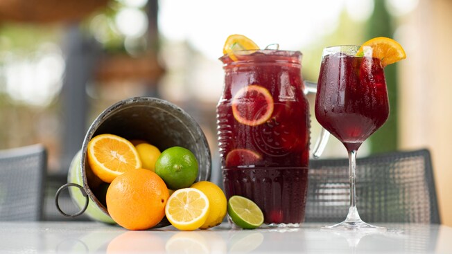 A glass of juice next to another container of juice and a bucket of lemons, oranges and limes