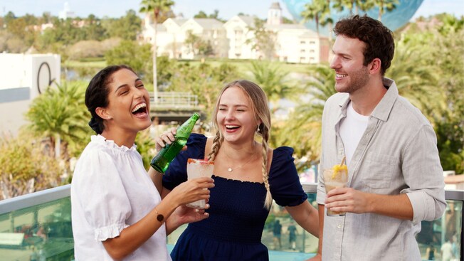 Three friends laugh while holding drinks on a rooftop bar