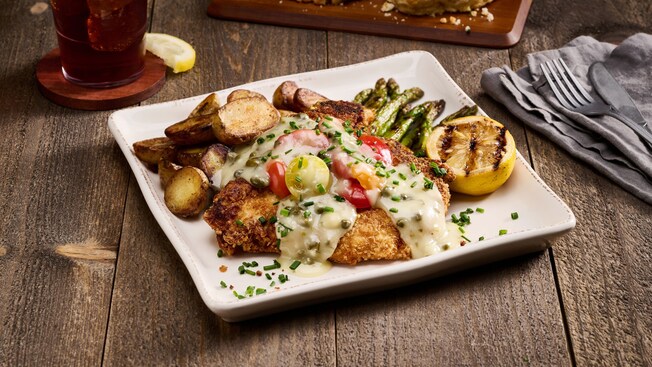 A dinner platter with a fried chicken cutlet topped with garlic, caper and gravy, served with potatoes and asparagus