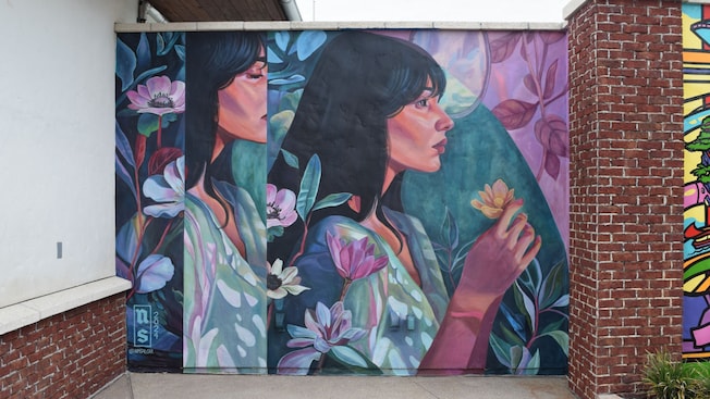 A mural against a brick wall by Nicole Salgar featuring 2 profiles of a woman surrounded by flowers and foliage, holding a flower