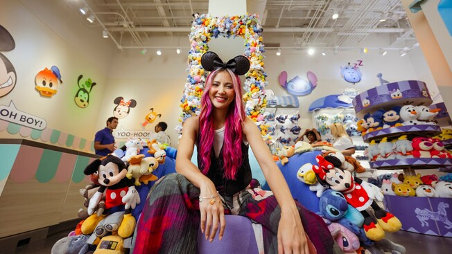 Una mujer con una diadema con orejas de Minnie Mouse sentada en un trono decorado con juguetes de peluche de Personajes de Disney dentro de la tienda Disney Ever After