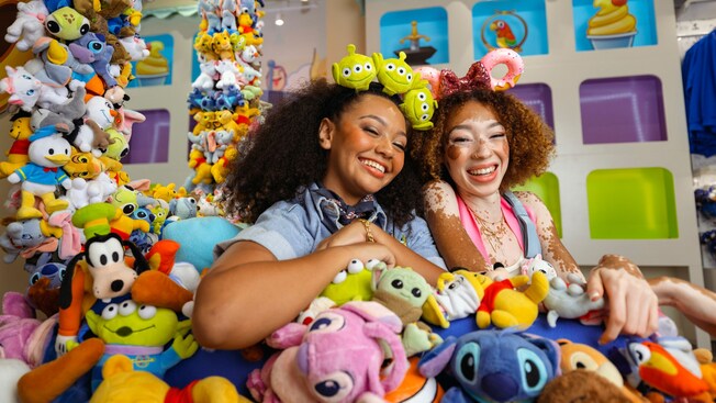 Dos mujeres con una diadema con temática de Toy Story y una diadema con orejas con temática de rosquilla de Minnie Mouse sentadas en un trono de juguetes de peluche de Disney dentro de la tienda Disney Ever After