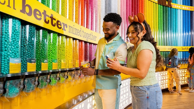 Un homme et une femme vêtus de vêtements sur le thème de Disney ramassent des M&M dans un magasin avec un mur de distributeurs M&M