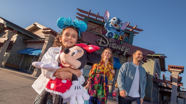 A girl hugging a large Minnie Mouse plush walking with her parents and a large Stitch figure above a sign with the words ‘World of Disney’ 