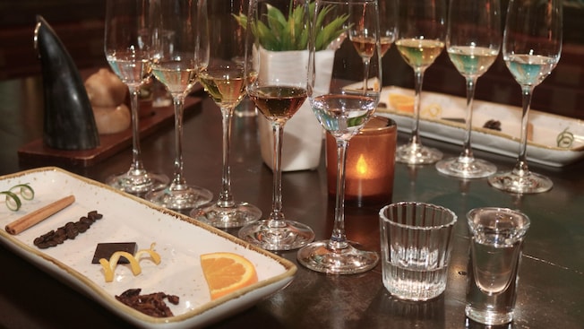 A table set for a tequila tasting, with 5 flute glasses, 2 shot glasses and a plate with a lime peel, a cinnamon stick, coffee beans, a block of chocolate, a lemon peel, an orange wedge and fried grasshoppers