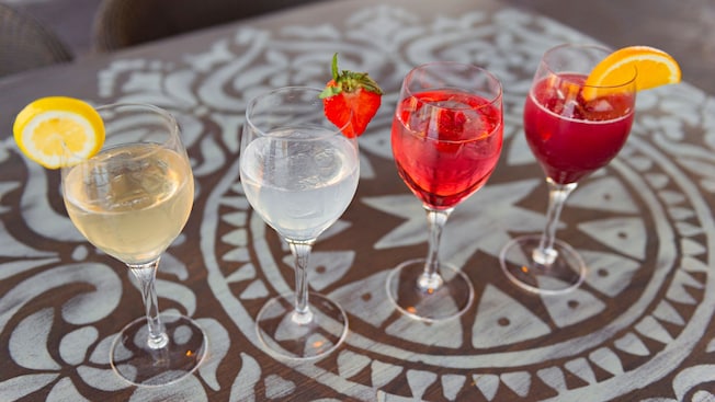 Four glasses, each holding a different type of sangria, are lined up in a row on a decorative table top