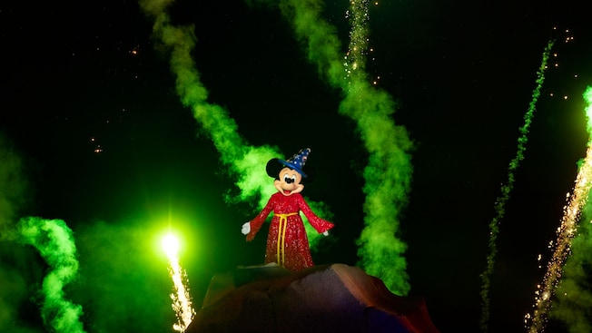 Sorcerer Mickey Mouse stands on top of a mountain while fireworks burst around him