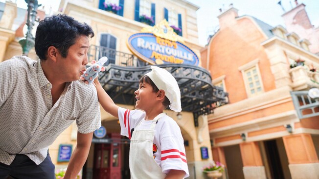 A young Guest holding a Remy plushie up to his father's shoulder in front of Remy's Ratatouille Adventure at Epcot