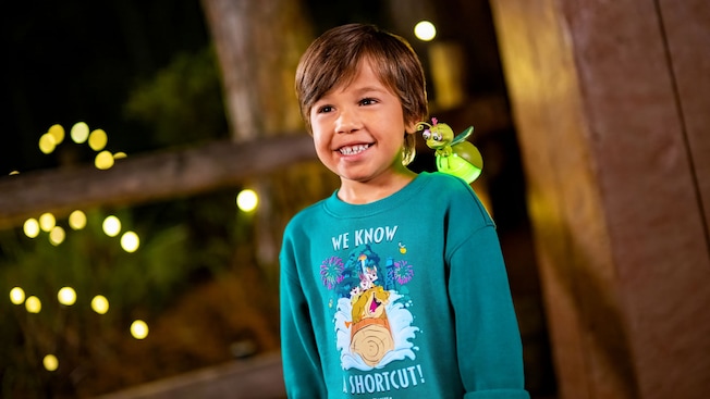 A smiling child wearing a Tiana’s Bayou Adventure sweatshirt with a cartoon firefly figurine perched on their shoulder