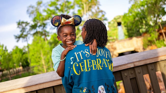 A smiling child wearing a Tiana’s Bayou Adventure Ear Headband hugging a woman wearing a Tiana’s Bayou Adventure jacket with the words, ‘It’s a joyful celebration’