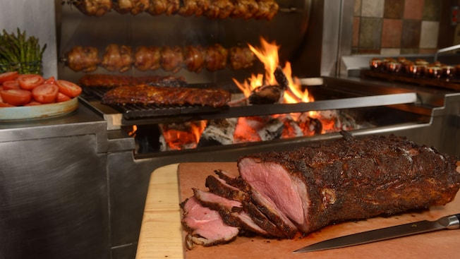 Vários frangos assados em estilo rotisserie, pedaços de carnes em uma grelha e um grande rosbife em uma tábua de cortes