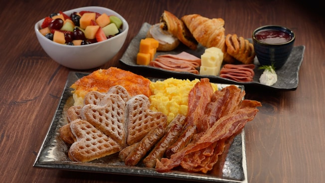 A platter of scrambled eggs, bacon, sausage, Norwegian waffles and potato casserole, a bowl of mixed fruit and a platter of pastries, cheeses and meats