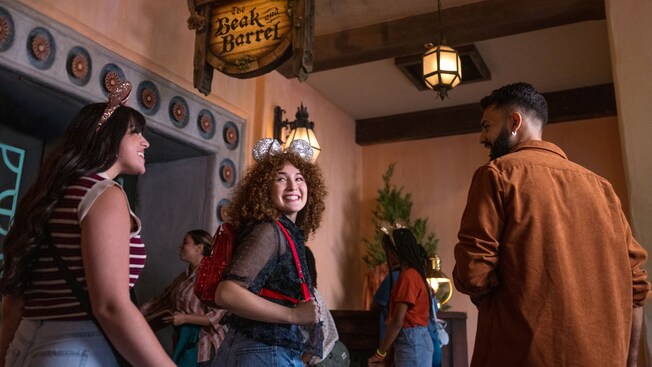 Adults in Mickey Ear headbands socialize beneath the sign for The Beak and Barrel at Magic Kingdom park