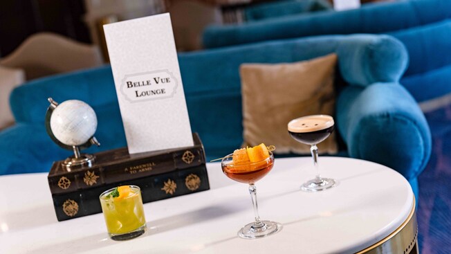 Uma mesa com 3 coquetéis e um cardápio escrito “Belle Vue Lounge” na frente do Disney’s Boardwalk Inn