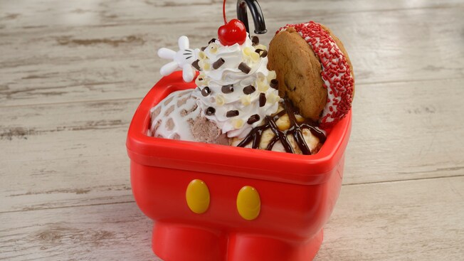 An ice cream sundae complete with whipped cream and a cookie sandwich served in a bowl shaped like Mickey Mouse’s pants