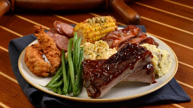 A plater of food that includes hickory smoked ribs, crispy Cajun chicken, smoked sausage, beef brisket, corn, mashed potatoes, green beans, and mac and cheese.