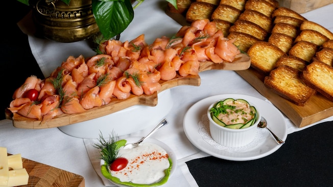 A table set with 3 wooden boards holding smoked salmon, toasted bread, slices of cheese, a plate with dipping sauce and a bowl with salmon spread