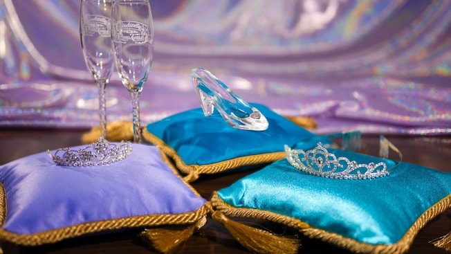 A Cinderella themed tiara, glass slipper and 2 champagne flutes each rest on 3 luxurious pillows