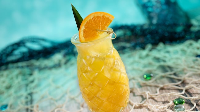 A tropical beverage in a pineapple shaped clear glass cup garnished with an orange slice