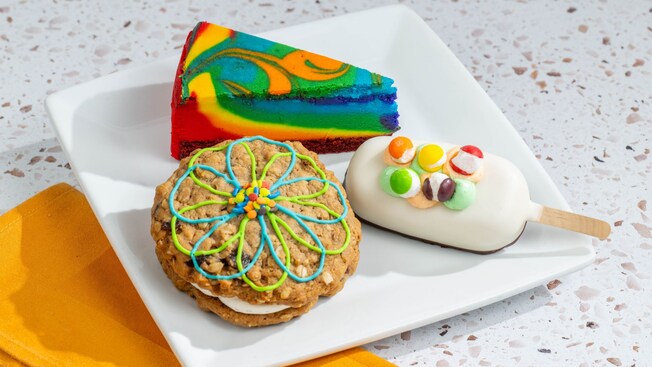 A plate featuring a slice of rainbow cheesecake next to a cake pop and a oatmeal cookie sandwich