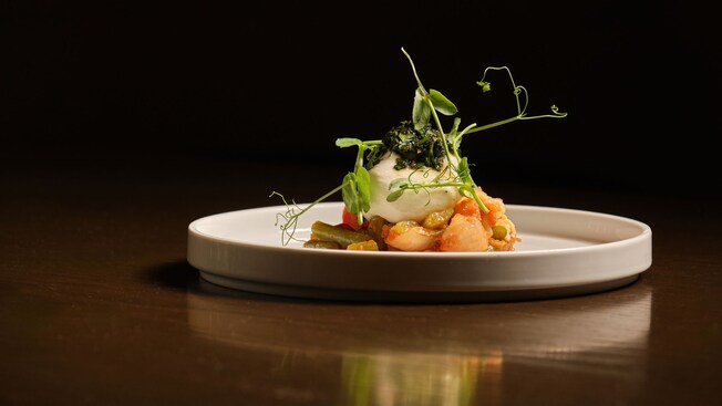 A plated dish of burrata topped with herbs and pea shoots, served over sautéed shrimp and vegetables