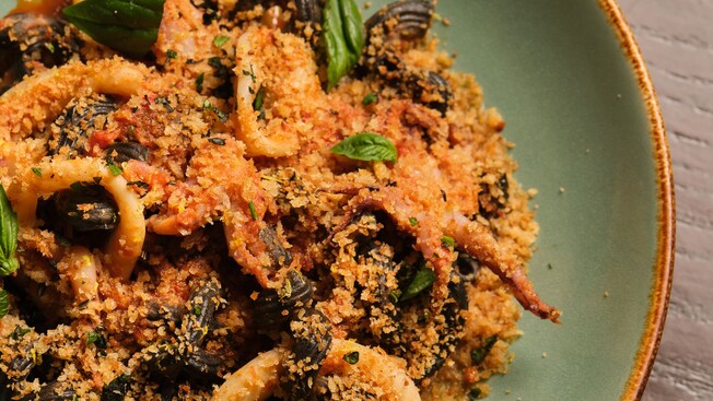 A ceramic bowl filled with squid ink pasta, calamari rings, mussels and baby octopus, topped with toasted breadcrumbs and basil leaves