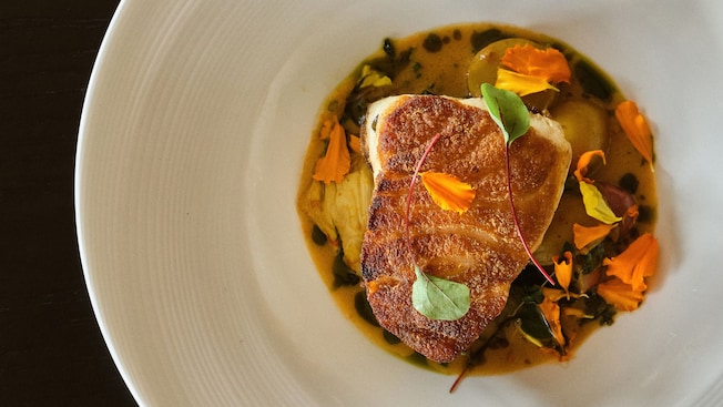 Un filete de pescado a la plancha, rodeado de salsa y decorado con flores comestibles y microvegetales