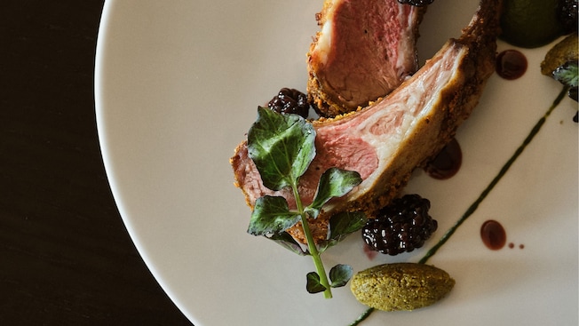 Dos chuletas de cordero en un plato, decoradas con moras, hierbas frescas y rociadas con salsa