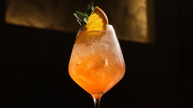 An iced cocktail in a stemmed wine glass garnished with an orange wedge and mint leaves