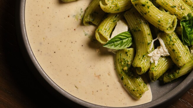 A ceramic dish of rigatoni pasta coated in pesto and topped with torn burrata and fresh basil leaves