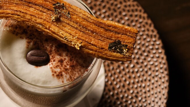 A glass filled with tiramisu, dusted with cocoa powder, topped with a single espresso bean and served with a churro resting across the rim