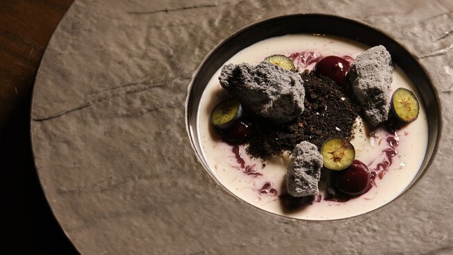 A bowl of panna cotta swirled with berry sauce, meringue pieces, chocolate crumble, whole cherries and sliced kumquats