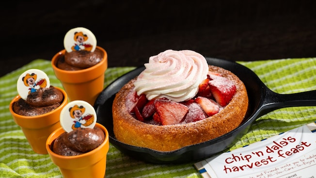 Un pastel pequeño con fresas y crema batida en una sartén, rodeado de 3 tazas de mousse de chocolate con figuras de Mickey Mouse como jardinero, cerca de un menú titulado ‘Chip and Dale’s Harvest Feast’