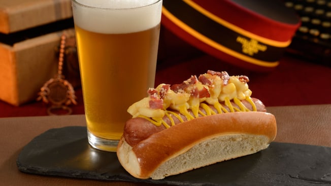 A hot dog sprinkled with mustard and with bacon and macaroni on top next sits to a glass of beer