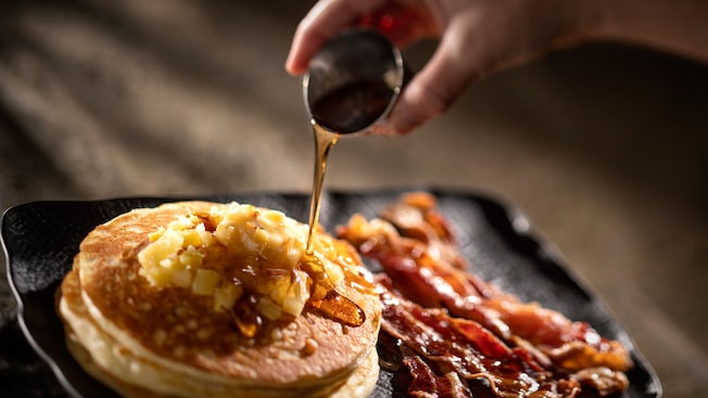A hand pouring a cup of syrup onto a plate of pancakes and bacon
