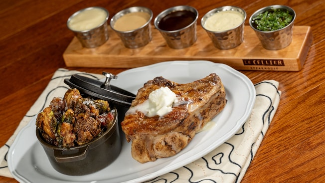 Une côtelette de porc Duroc grillée et vieillie à sec de 14 onces (397 g) avec des choux de Bruxelles rôtis, servie dans une assiette accompagnée d’un assortiment de 5 sauces du restaurant Le Cellier Steakhouse