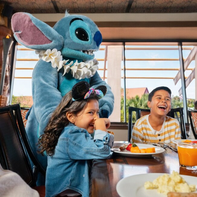 Two children and their grandfather at a table talking to Stitch and having breakfast at 'Ohana