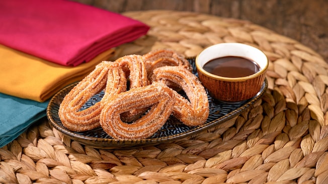 A plate of cinnamon sugar Churro Loops served with a cup of chocolate dipping sauce on a woven placemat