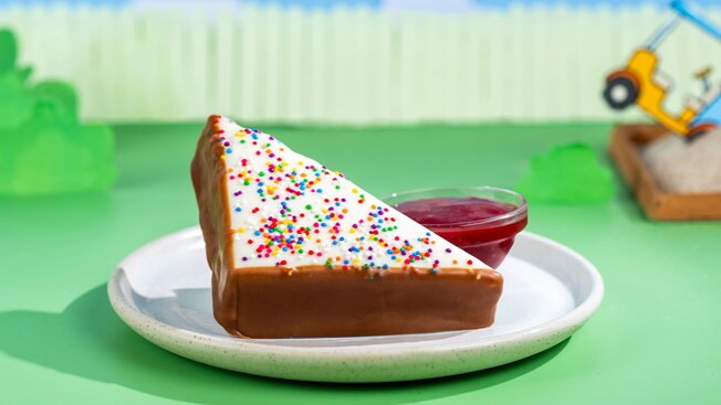 Slice of Fairy Bread Cake dipped in white chocolate and rainbow sprinkles with a side of raspberry sauce