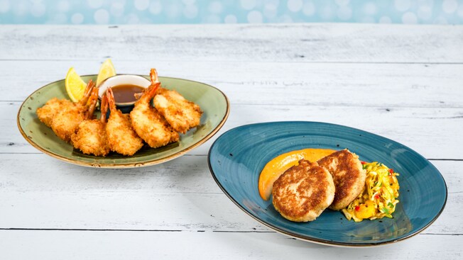 A plate with coconut shrimp, lemon wedges and dipping sauce next to a plate with 2 crab cakes, sauce and corn salad from Sebastian's Bistro