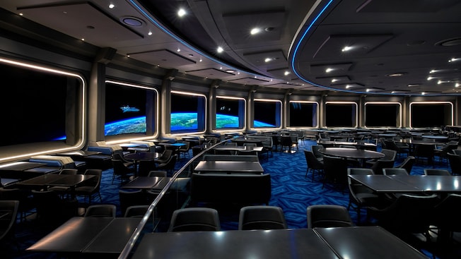 An image of an empty restaurant with black tables and chairs themed to look like a space station just above the planet Earth.