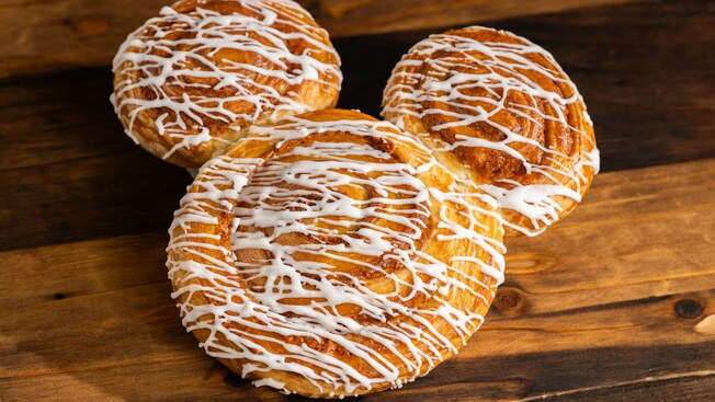 A Mickey Mouse shaped Cinnamon Roll drizzled with white icing on a wooden surface